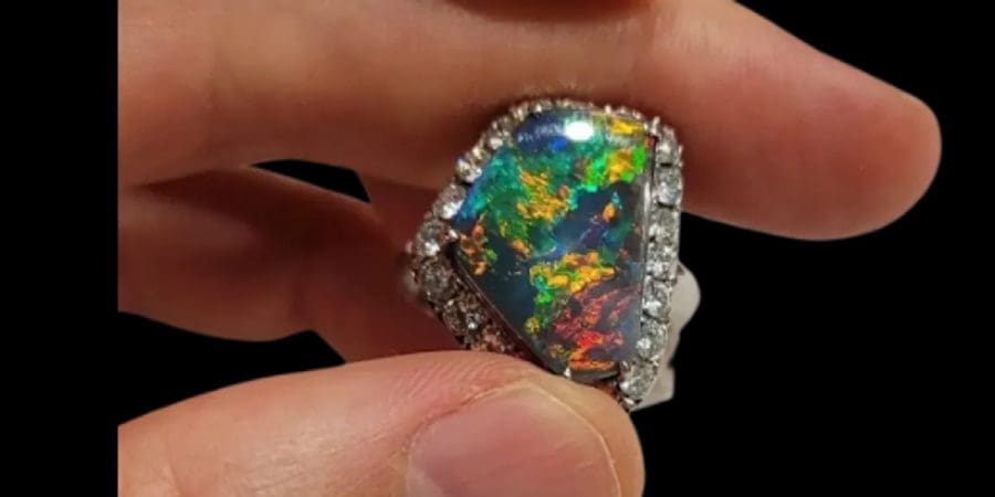 A hand holding a large black Australian opal ring surrounded by diamonds, with the opal displaying vibrant multicoloured patterns.