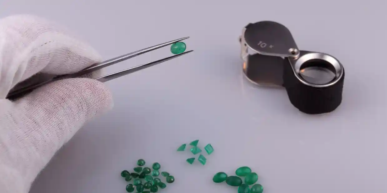 A gloved hand holding tweezers with a green gemstone next to a pile of similar stones and a jeweler's loupe on a white surface.