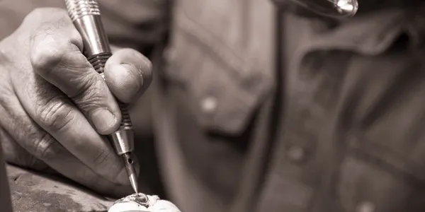 Close-up of a person's hand using a soldering iron on a small metal object, depicted in black and white.