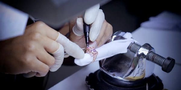 A jeweler in gloves uses a tool to intricately work on a piece of jewelry secured in a clamp.